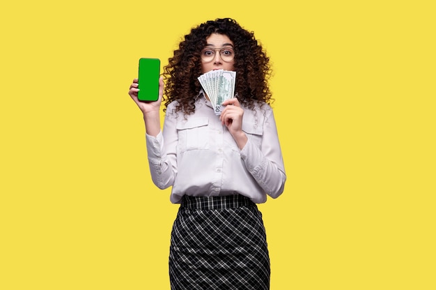 La donna sorpresa in vetri tiene la pila di dollari e smartphone con lo schermo verde in bianco sopra fondo isolato giallo. Concetto di casinò online, scommesse, giochi