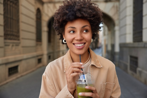 La donna sorride ampiamente beve rinfrescante frullato verde dalla paglia passeggiate in città durante il tempo libero vestita in giacca marrone pone sulla città antica
