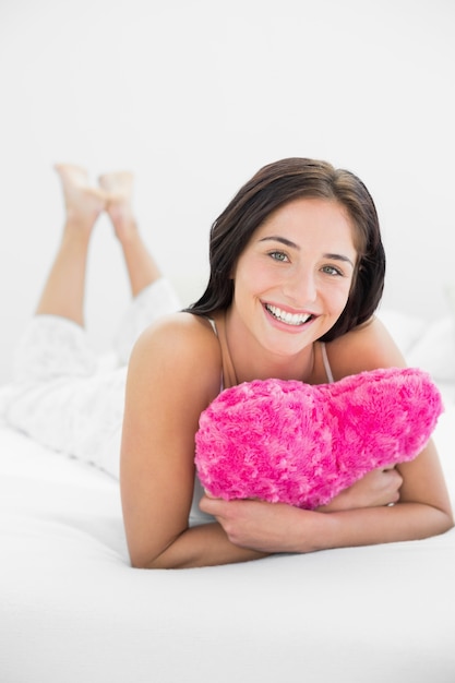 La donna sorridente che si trova con il cuore ha modellato il cuscino a letto