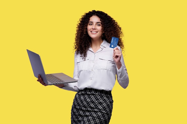 La donna sorridente con capelli ricci tiene il computer portatile e la carta di credito sopra gli sfondi isolati gialli