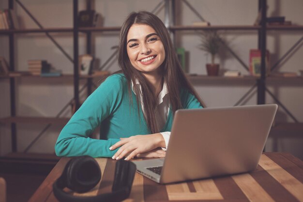 La donna sorridente con le cuffie è seduta al tavolo di legno con il computer portatile sul tavolo