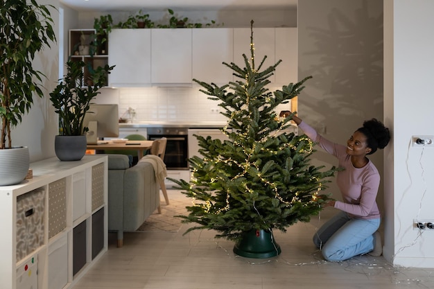 La donna sorridente decora l'albero di natale con luci a ghirlanda a led si gode il capodanno e i preparativi per il natale