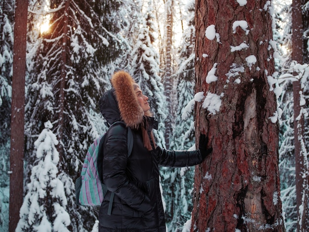 La donna sorridente felice in vestiti caldi con un cappuccio sulla sua testa tocca la corteccia dell'albero. Unità con la natura. Bella pineta sotto la neve, gelido inverno.