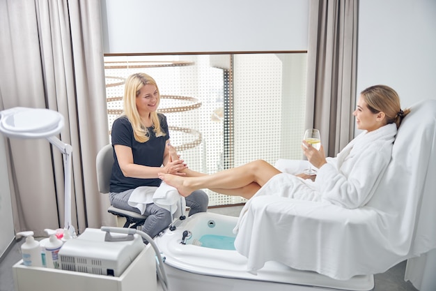 La donna sorridente si sta godendo il massaggio ai piedi di una signora professionista mentre beve un bicchiere di acqua gelidaing