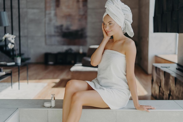 La donna sottile applica la crema per il viso in un moderno asciugamano sulla testa Concetto di bellezza e cura della pelle