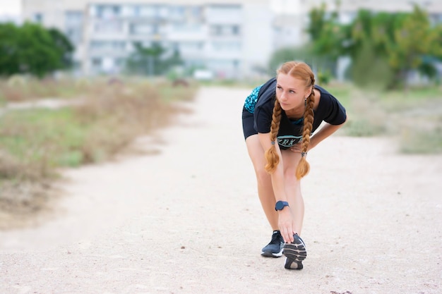 La donna sportiva dai capelli rossi tirando la gamba che si prepara per l'allenamento distoglie lo sguardo sulla strada