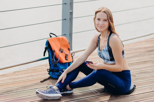 La donna sportiva indossa leggings casual e scarpe sportive guarda positivamente la fotocamera tiene il cellulare in mano ascolta la radio online con gli auricolari si rompe dopo l'allenamento all'aperto Concetto di passatempo