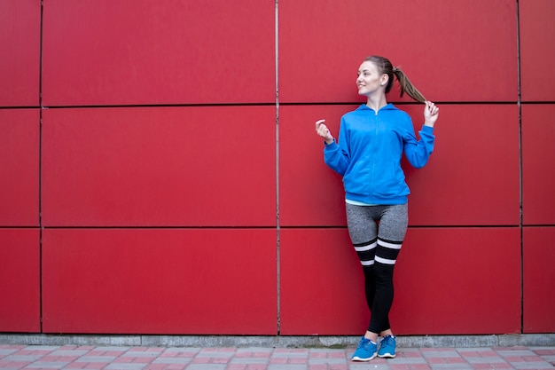 La donna sportiva sta contro una parete rossa in abiti sportivi, la donna di forma fisica che posa su una strada cittadina, copia spazio