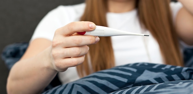 La donna sta controllando la sua temperatura sdraiata sul letto a guardare il termometro