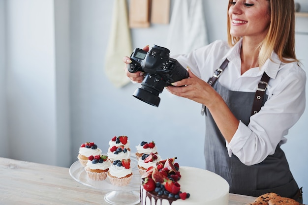 La donna sta in cucina e scatta una foto dei suoi biscotti e della sua torta fatti in casa usando la fotocamera.