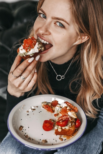 La donna sta mangiando un gustoso toast di avocado con pomodori, salmone e formaggio da un piatto.