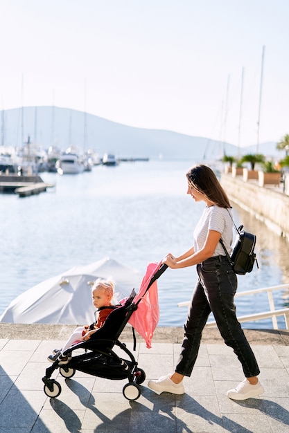 La donna sta spingendo sua figlia in un passeggino rosa su un molo in una giornata di sole.