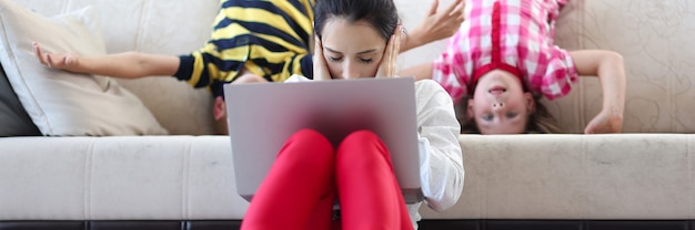 La donna stanca con il computer portatile si siede sul pavimento con le orecchie dei palmi chiuse dietro sul divano i bambini si concedono