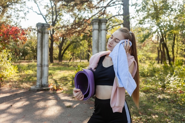 La donna stanca in forma pulisce il sudore dal viso con un asciugamano e tiene il tappetino da yoga all'aperto