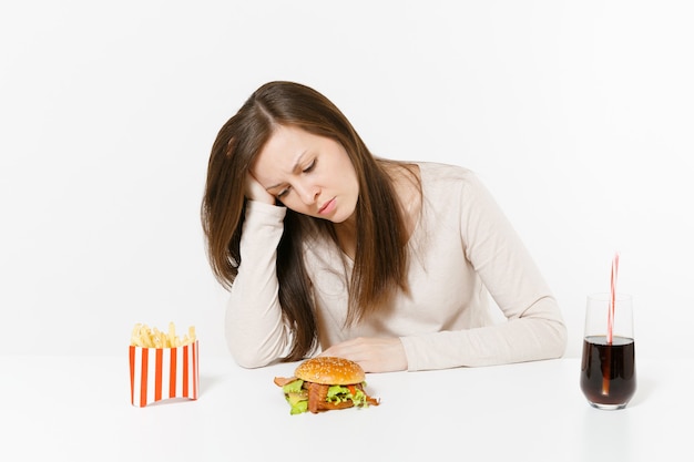 La donna stanca triste ha messo la mano sulla testa che si siede alla tavola con l'hamburger, le patate fritte, la cola in bottiglia di vetro isolata su fondo bianco. Una corretta alimentazione o un classico fast food americano. Area con copia spazio.