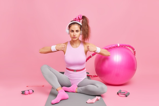 la donna stringe il pugno dimostra il suo potere vestita in abbigliamento sportivo ha una coda di cavallo ascolta musica tramite le cuffie utilizza attrezzature per ginnastica pilates. Allenamento sportivo e stile di vita sano