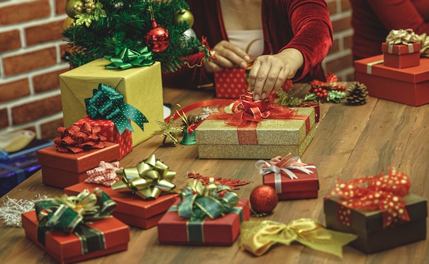 La donna sul maglione mette una lussuosa scatola regalo dorata con splendido fiocco sul tavolo di bellissimi ornamenti e nastro colorato per preparare un regalo meraviglioso per la decorazione della festa a casa del festival delle vacanze di Natale