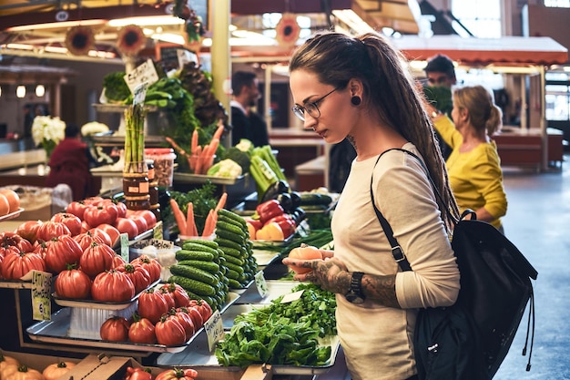 La donna tatuata pensosa sceglie con cura pomodori e altre verdure su un mercato locale.