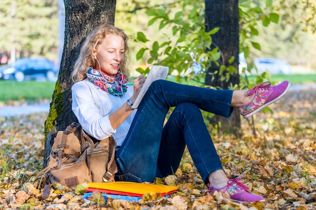 La donna teenager dello studente che si siede sul prato inglese in parco con i libri e taccuino e scrive