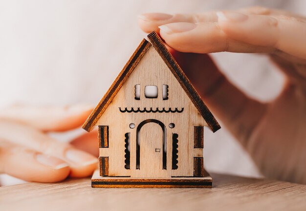 La donna tiene e protegge una casa in legno con le mani con il sole su uno sfondo rosa chiaro. Dolce casa