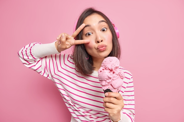 la donna tiene le labbra piegate fa un gesto di pace sugli occhi mangia un delizioso gelato ha un debole per i dolci sciocchezze in giro indossa cuffie wireless ascolta musica vestita con un maglione a righe.