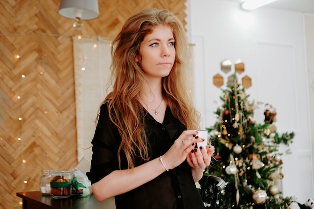 La donna tiene una tazza di caffè al mattino nella cucina di Natale. Felice giovane donna che si diverte e sorride