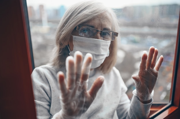 La donna triste anziana senior sola in maschera medica e guanti che esaminano la finestra rimangono l'isolamento a casa per l'auto quarantena. Concetto di quarantena domestica, prevenzione COVID-19, situazione di epidemia di coronavirus