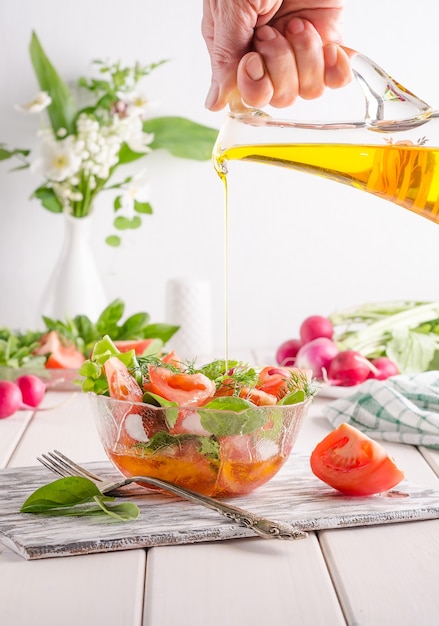 La donna versa l'olio in un'insalata con verdure ed erbe aromatiche in un'insalatiera trasparente su una superficie di legno bianca. Mangiare sano