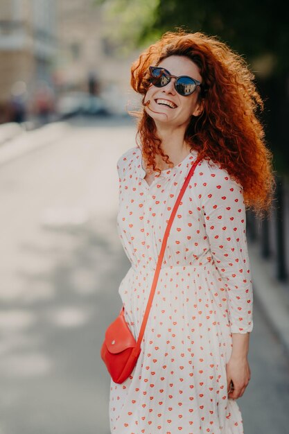 La donna volpe ottimista con un'espressione felice guarda in lontananza indossa sfumature abito lungo passeggiate all'aperto in strada gode di una bella giornata estiva ha un ampio sorriso Stile di vita delle persone riposo e stile di vita