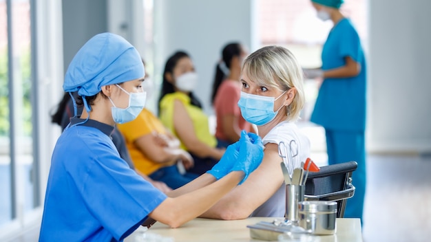 La dottoressa asiatica indossa la maschera facciale e l'uniforme ospedaliera blu che inietta il vaccino a una donna anziana caucasica alla scrivania del reparto con attrezzature mentre altri pazienti aspettano in coda su sfondo sfocato.