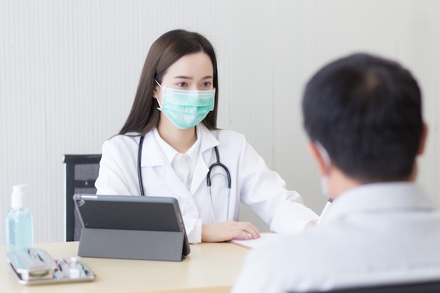 La dottoressa asiatica indossa una maschera medica e registra i sintomi di un paziente uomo