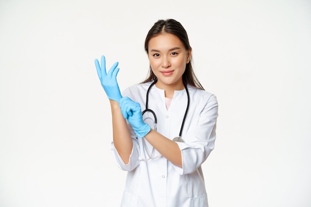 La dottoressa asiatica sorridente e sicura ha indossato guanti medici, preparandosi per l'esame del paziente, in piedi in uniforme su sfondo bianco.