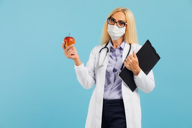 La dottoressa matura con gli occhiali tiene la mela e consiglia di mangiare acqua di frutta su sfondo blu. Dieta e concetto di perdita di peso