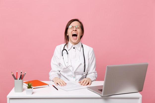 La dottoressa pazza stanca si siede alla scrivania al computer con documento medico in ospedale isolato su sfondo rosa pastello. Donna in abito medico occhiali stetoscopio. Concetto di medicina sanitaria.