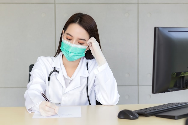 La dottoressa professionista asiatica lavora seriamente mentre scrive il documento di trattamento del paziente. Indossa una maschera medica e guanti in lattice nella sala degli esami clinici dell'ospedale nel concetto di assistenza sanitaria
