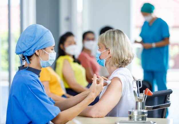 La dottoressa professionista indossa la maschera facciale e l'uniforme ospedaliera blu che inietta l'ago della siringa del vaccino contro il coronovirus nella spalla del paziente della donna anziana caucasica alla scrivania del reparto della clinica.