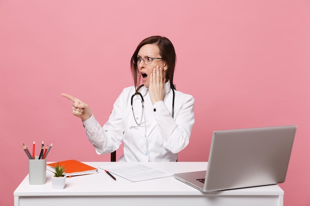 La dottoressa scioccata si siede alla scrivania al lavoro sul computer con documento medico in ospedale isolato su sfondo rosa pastello. Donna in abito medico occhiali stetoscopio. Concetto di medicina sanitaria.