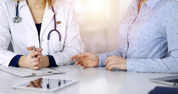 La dottoressa sconosciuta sta ascoltando il suo paziente, mentre è seduta insieme alla scrivania nell'armadietto soleggiato di una clinica. Il medico femminile con uno stetoscopio sta scrivendo negli appunti, da vicino. Perfetto m