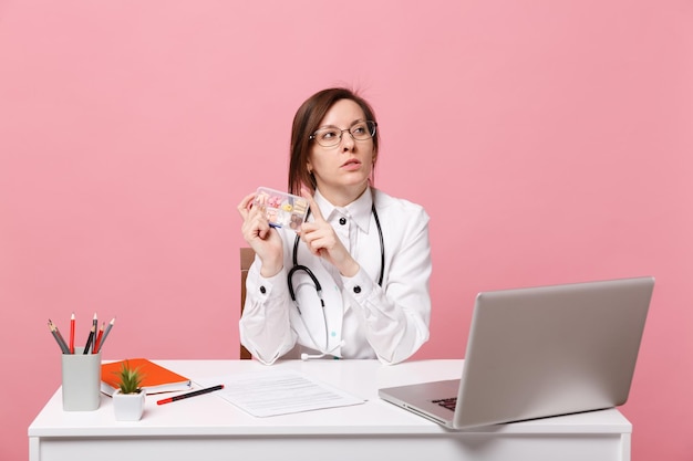 La dottoressa si siede alla scrivania al lavoro sul computer con le pillole della tenuta del documento medico in ospedale isolato sul fondo della parete rosa pastello. Donna in abito medico occhiali stetoscopio. Concetto di medicina sanitaria.