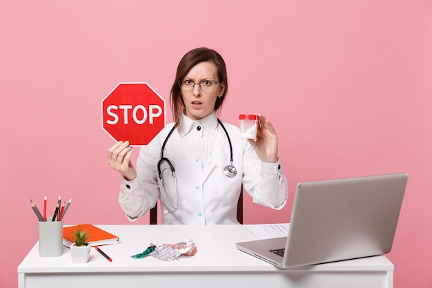 La dottoressa si siede alla scrivania al lavoro sul computer con le pillole della tenuta del documento medico in ospedale isolato sul fondo della parete rosa pastello. Donna in abito medico occhiali stetoscopio. Concetto di medicina sanitaria.