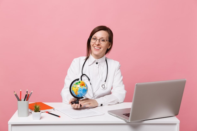 La dottoressa si siede alla scrivania al lavoro sul computer con un documento medico tiene il globo in ospedale isolato su sfondo rosa pastello. Donna in abito medico occhiali stetoscopio. Concetto di medicina sanitaria.