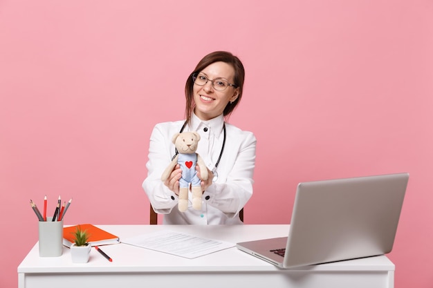 La dottoressa si siede alla scrivania e lavora al computer con il giocattolo della tenuta del documento medico in ospedale isolato su sfondo rosa pastello. Donna in abito medico occhiali stetoscopio. Concetto di medicina sanitaria.
