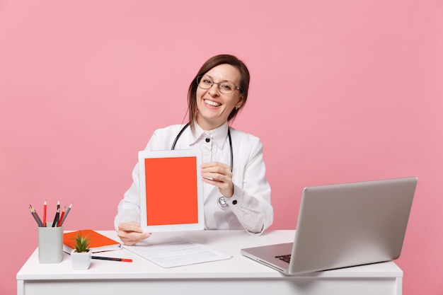 La dottoressa si siede alla scrivania e lavora al computer con il tablet della tenuta del documento medico in ospedale isolato su sfondo rosa pastello. Donna in abito medico occhiali stetoscopio. Concetto di medicina sanitaria