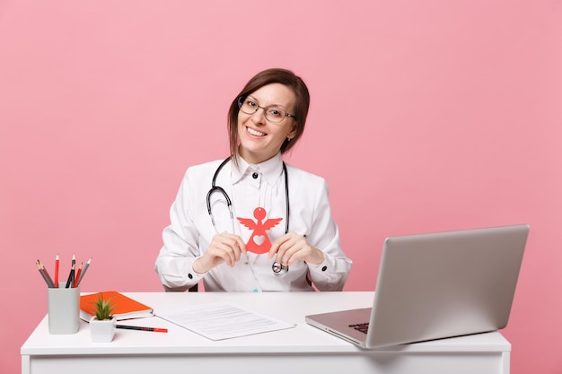 La dottoressa si siede alla scrivania e lavora al computer con un documento medico che tiene l'angelo in ospedale isolato su sfondo rosa pastello. Donna in abito medico occhiali stetoscopio. Concetto di medicina sanitaria.