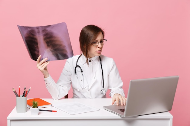 La dottoressa si siede alla scrivania e lavora al computer con un documento medico tiene i raggi x in ospedale isolato su sfondo rosa pastello. Donna in abito medico occhiali stetoscopio. Concetto di medicina sanitaria.