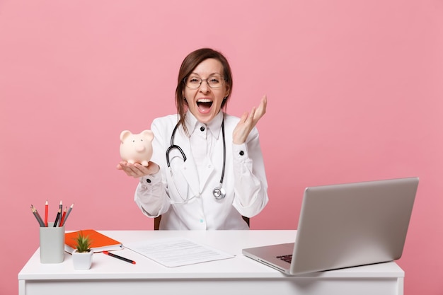 La dottoressa si siede alla scrivania e lavora al computer con un documento medico tiene i soldi del maiale in ospedale isolato su sfondo rosa pastello. Donna in abito medico occhiali stetoscopio. Concetto di medicina sanitaria.