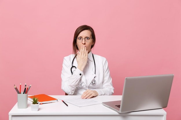 La dottoressa triste e stanca si siede alla scrivania al computer con documento medico in ospedale isolato su sfondo rosa pastello. Donna in abito medico occhiali stetoscopio. Concetto di medicina sanitaria.