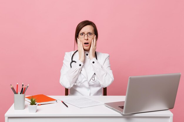 La dottoressa triste e stanca si siede alla scrivania al computer con documento medico in ospedale isolato su sfondo rosa pastello. Donna in abito medico occhiali stetoscopio. Concetto di medicina sanitaria.