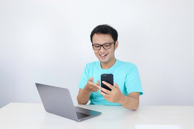 La faccia felice del giovane uomo asiatico durante la videochiamata o cosa al telefono con il laptop accanto a uno sfondo grigio isolato Uomo indonesiano che indossa una camicia blu