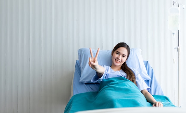 La faccina sorridente di una giovane donna asiatica solleva due dita per combattere con la malattia sul letto sullo sfondo dell'ospedale della stanza.